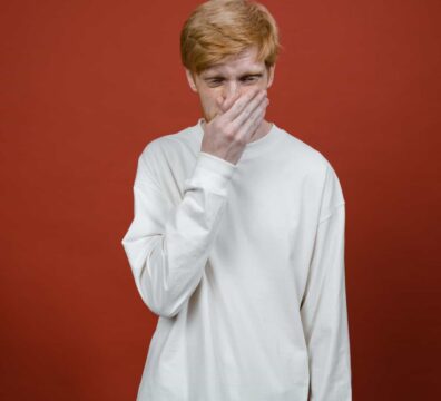 Young man in white sweater with hand on mouth standing against a red studio background.